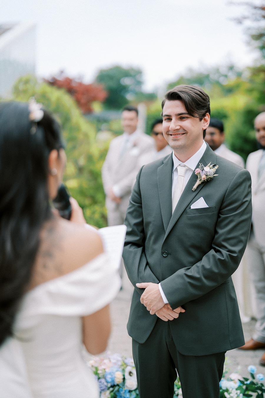 A groom in a formal suit smiling during an outdoor wedding ceremony, with the bride reading vows and groomsmen in the background. Lush greenery surrounds the venue, enhancing the romantic atmosphere.