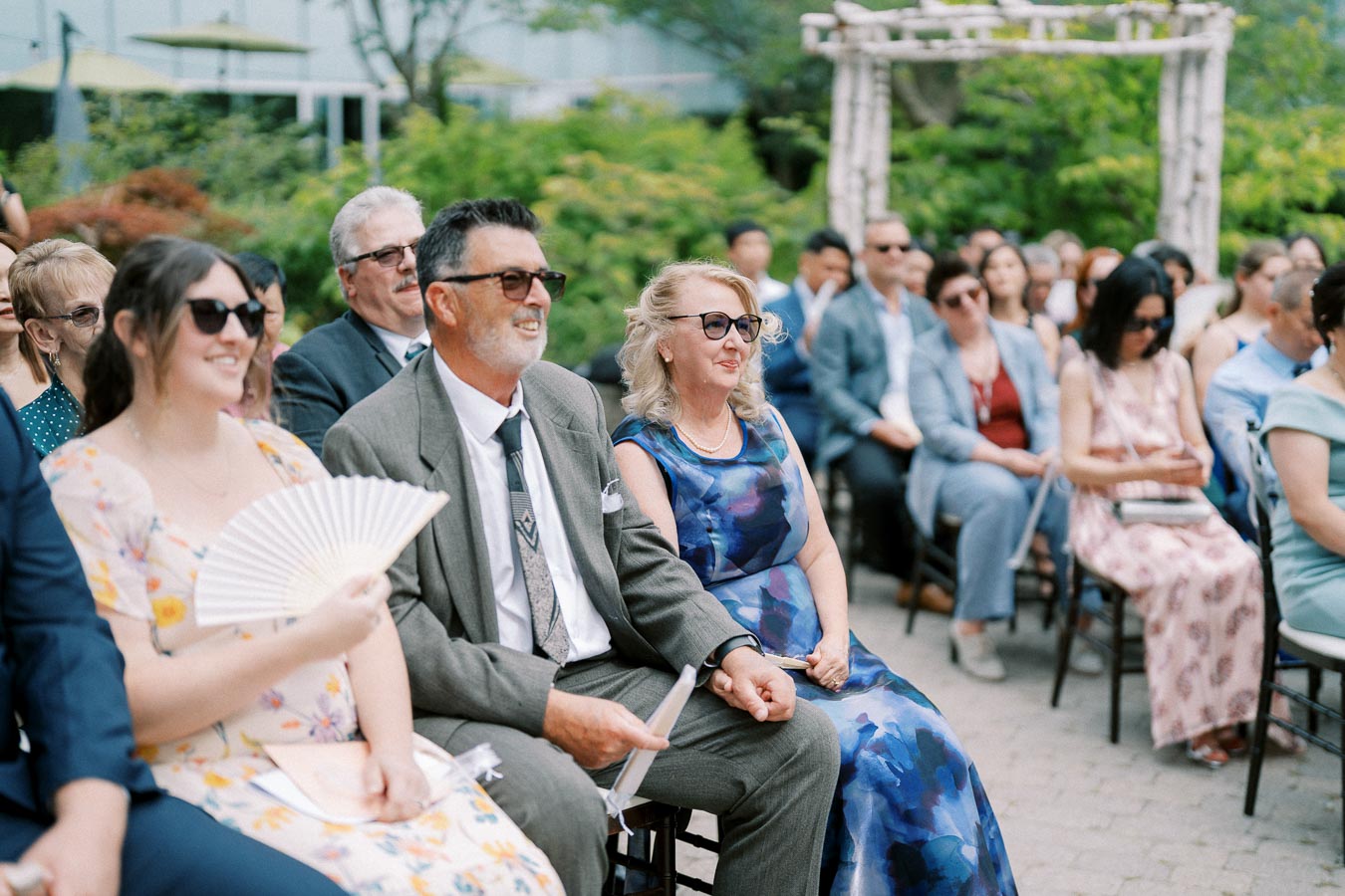 Outdoor wedding ceremony with guests seated in formal attire, enjoying the event on a sunny day with lush greenery in the background.