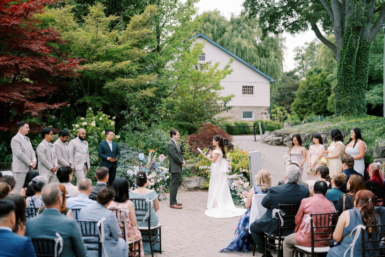 Outdoor garden wedding ceremony with a bride in a white dress reading vows to the groom, surrounded by bridesmaids and groomsmen, with guests seated in the foreground. The lush greenery and colorful flowers create a picturesque setting.