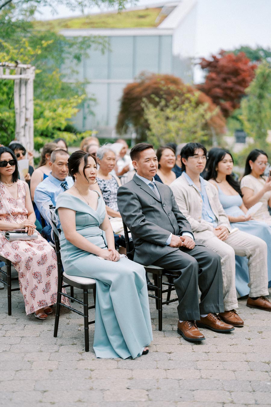Group of elegantly dressed guests sitting outdoor at a wedding ceremony, with lush greenery and modern architecture in the background.