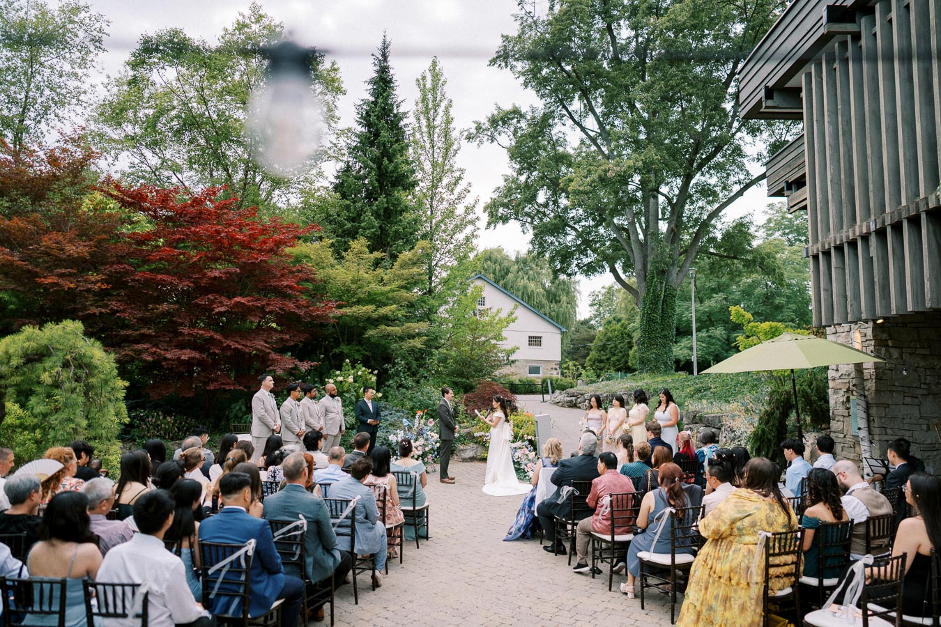 Outdoor wedding ceremony in a lush garden setting, featuring a bride and groom exchanging vows surrounded by guests and vibrant greenery.