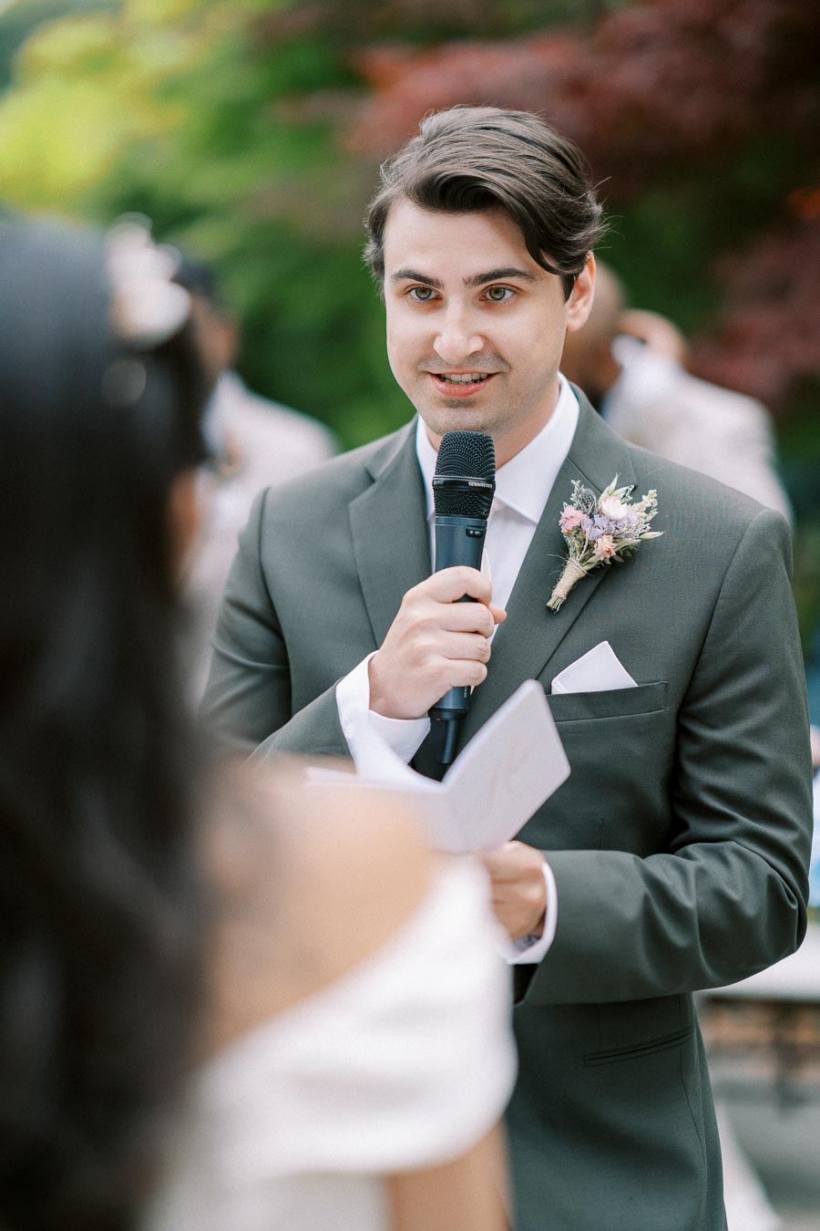 Groom in a green suit holding a microphone, giving a speech outdoors during a wedding ceremony.