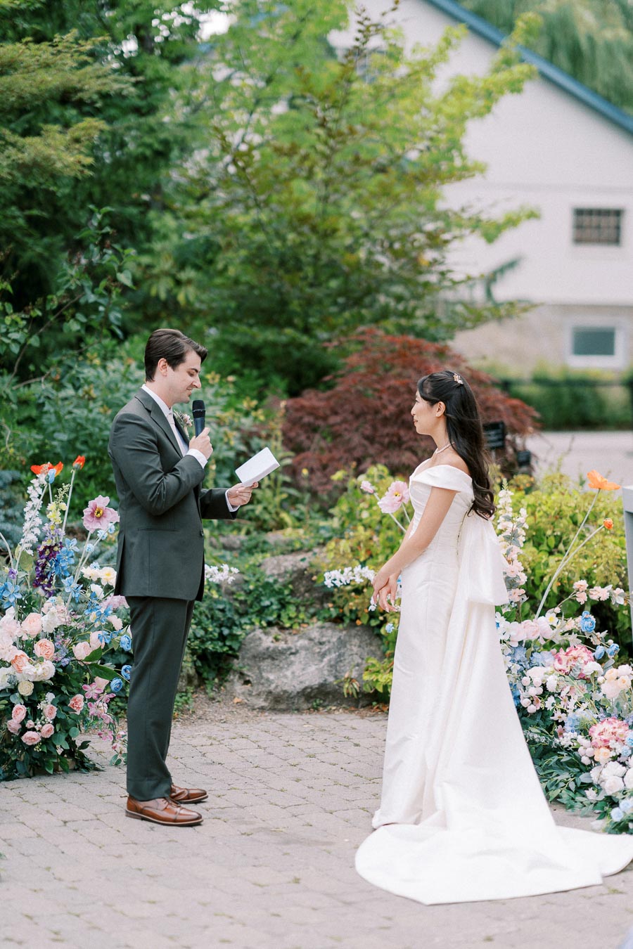A bride and groom exchange vows in a lush garden setting, surrounded by colorful flowers and greenery, with the groom holding a microphone and the bride wearing an elegant white gown.