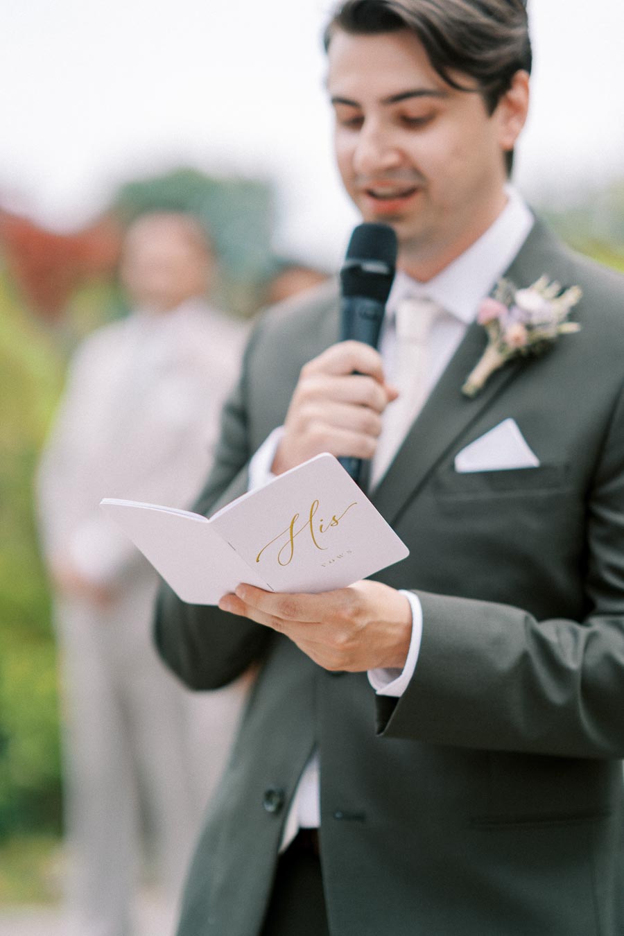 A groom in a suit reads wedding vows from a booklet labeled His Vows while holding a microphone during an outdoor ceremony.