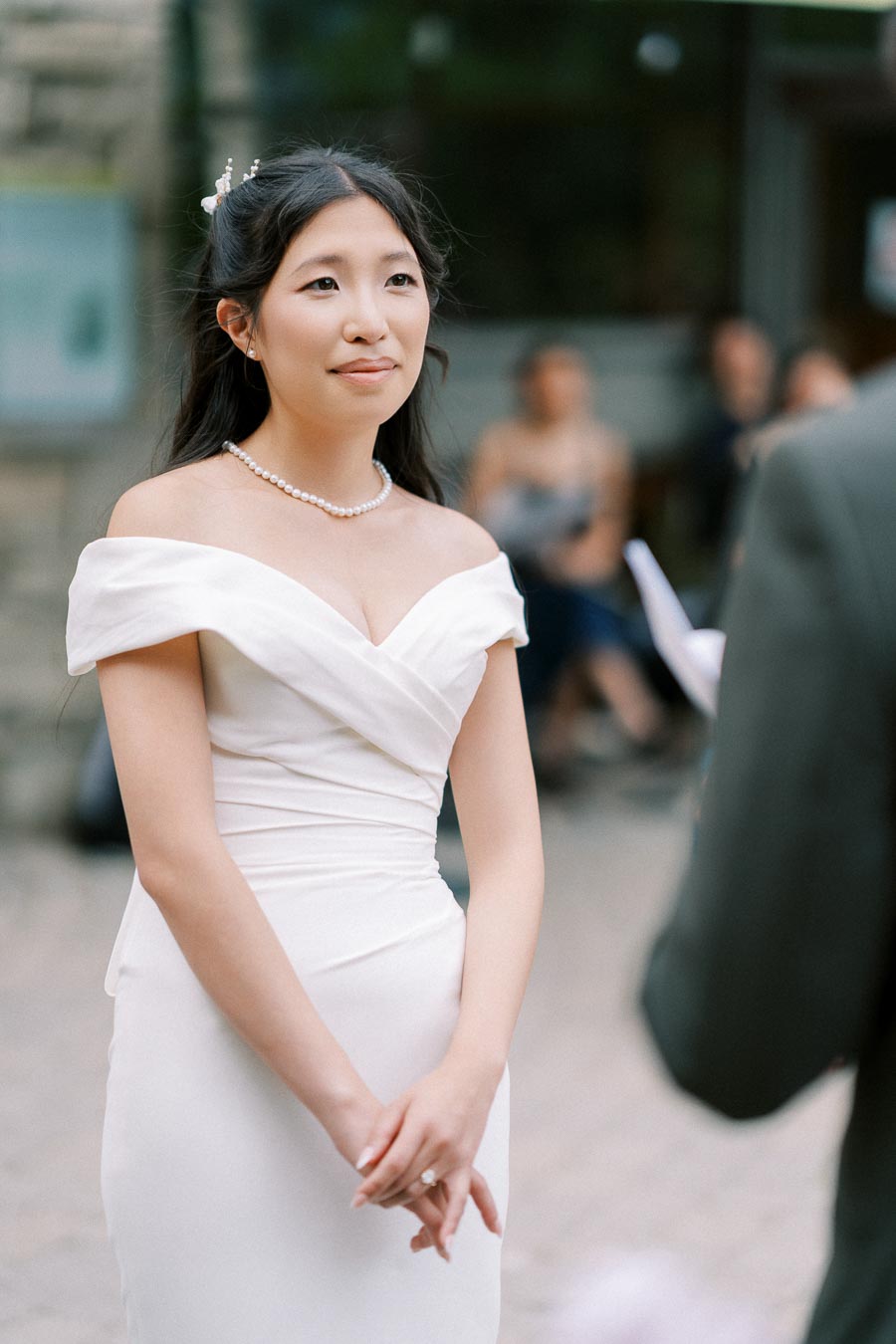 A bride in an elegant off-the-shoulder wedding dress stands during an outdoor ceremony, wearing a pearl necklace and styled hair, with blurred guests in the background.