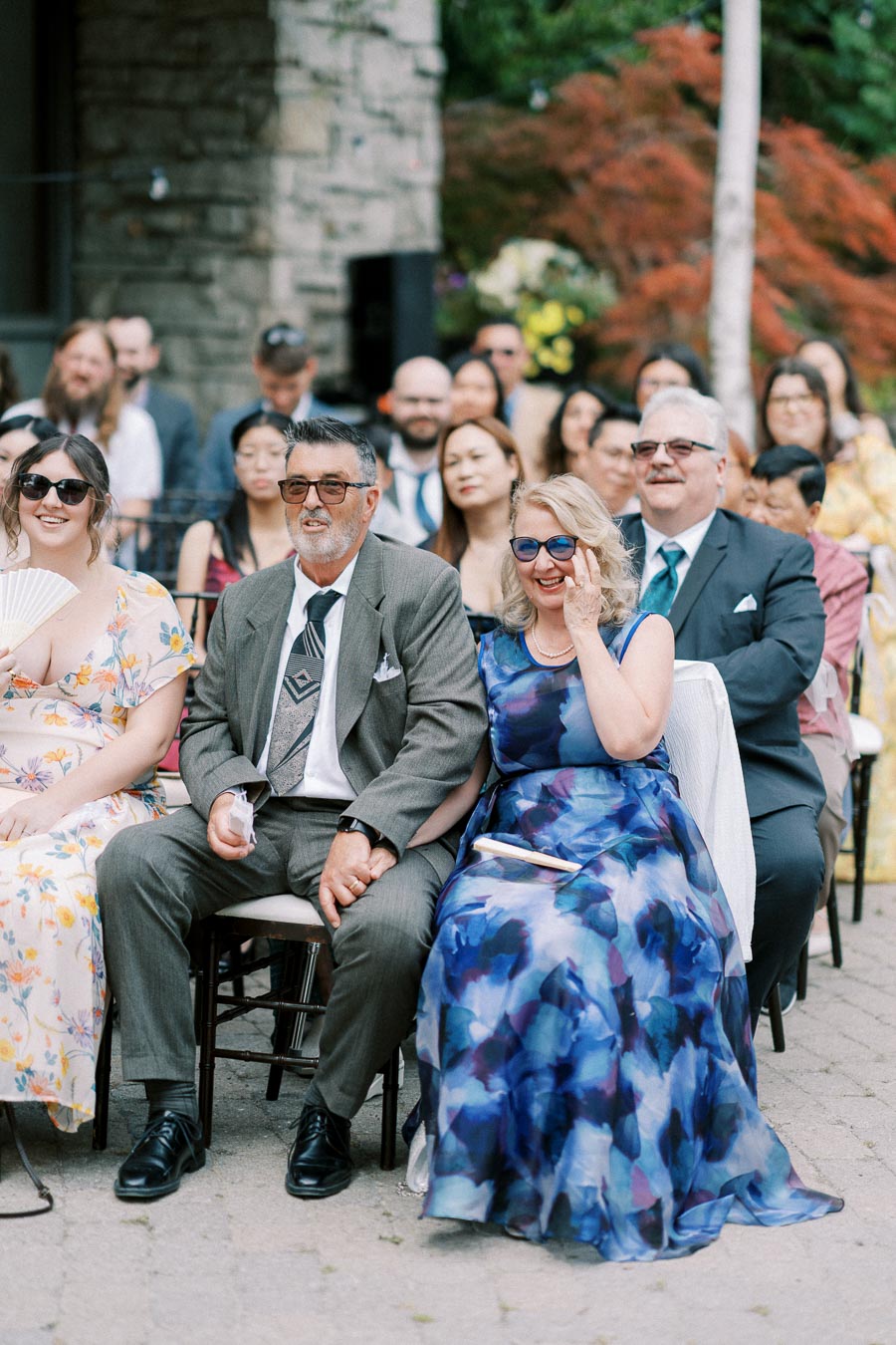Audience seated at an outdoor wedding ceremony, featuring women in colorful dresses and men in suits, with a stone building and lush greenery in the background.