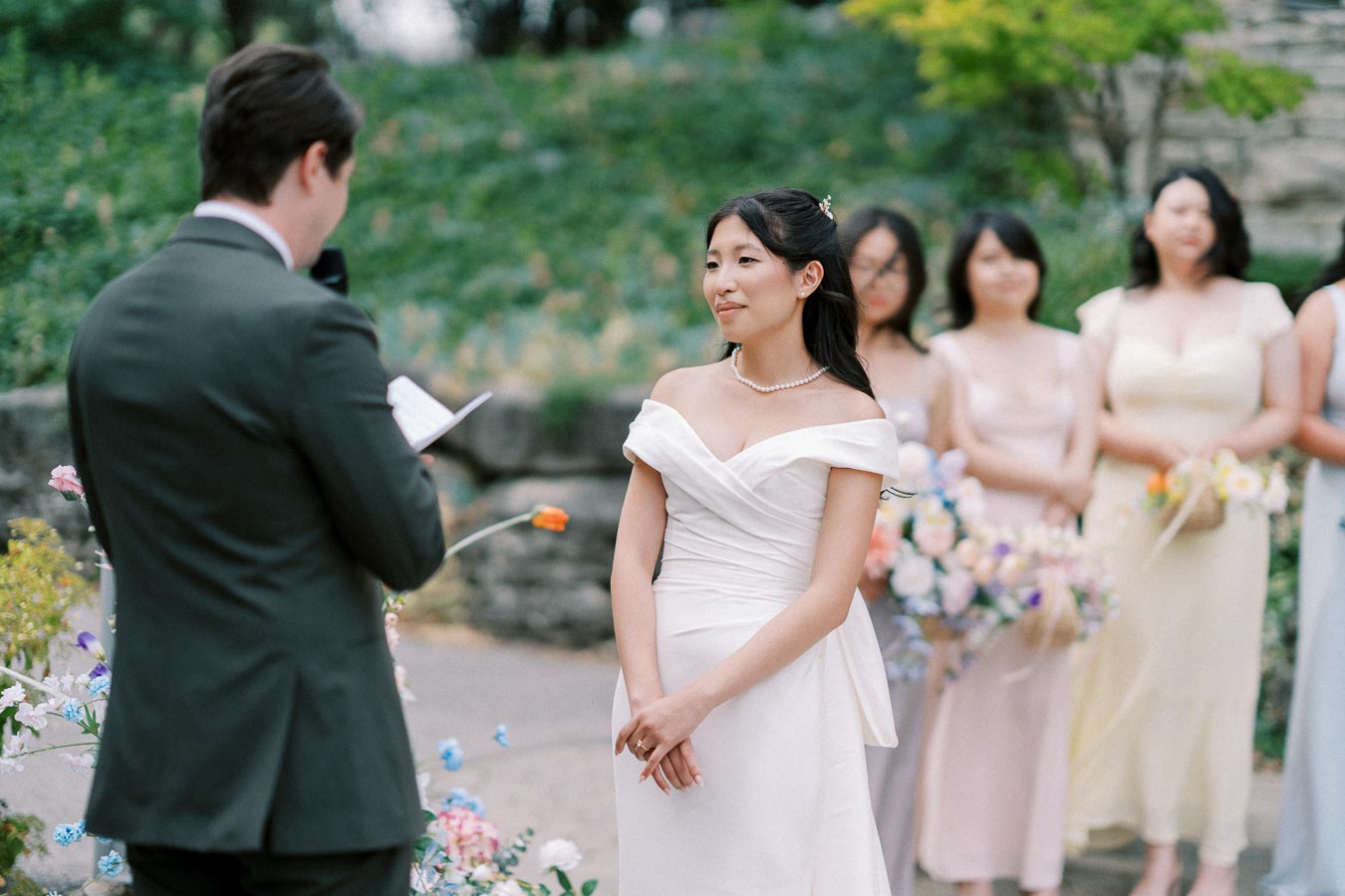A bride in an elegant white gown stands facing the groom during an outdoor wedding ceremony, surrounded by bridesmaids holding colorful bouquets, with a lush green background.