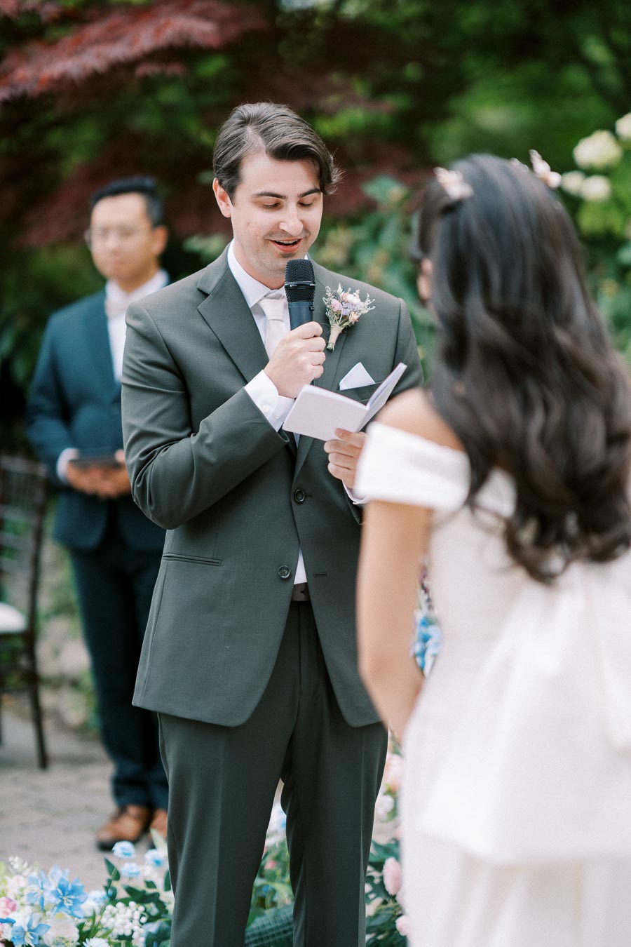 Groom reading vows to bride during outdoor wedding ceremony, with guests and floral decorations in the background.