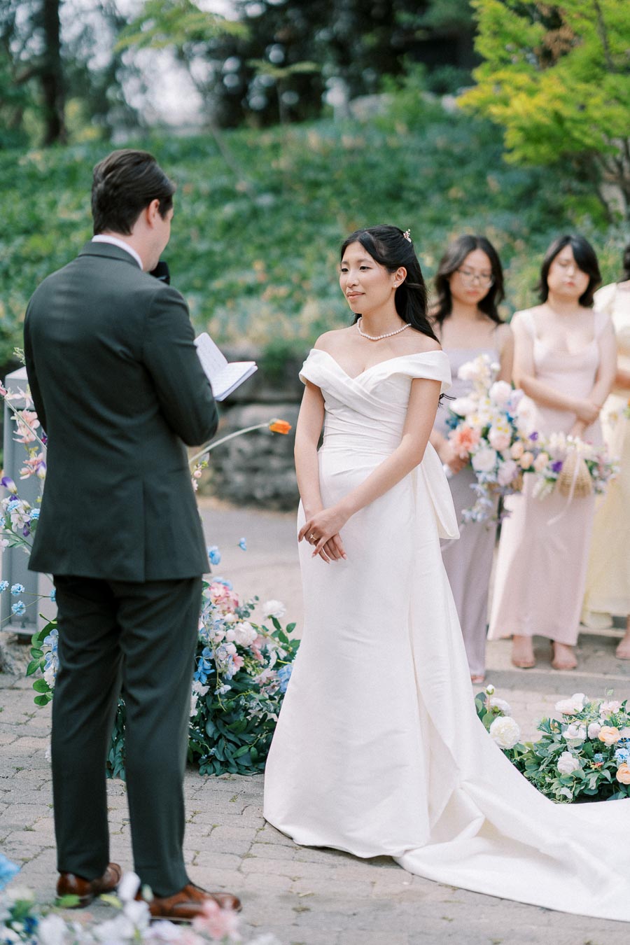 A bride in an elegant white gown stands in an outdoor wedding ceremony, listening to the groom who is reading vows from a book. Bridesmaids in soft pastel dresses hold floral bouquets in the background, surrounded by lush greenery.