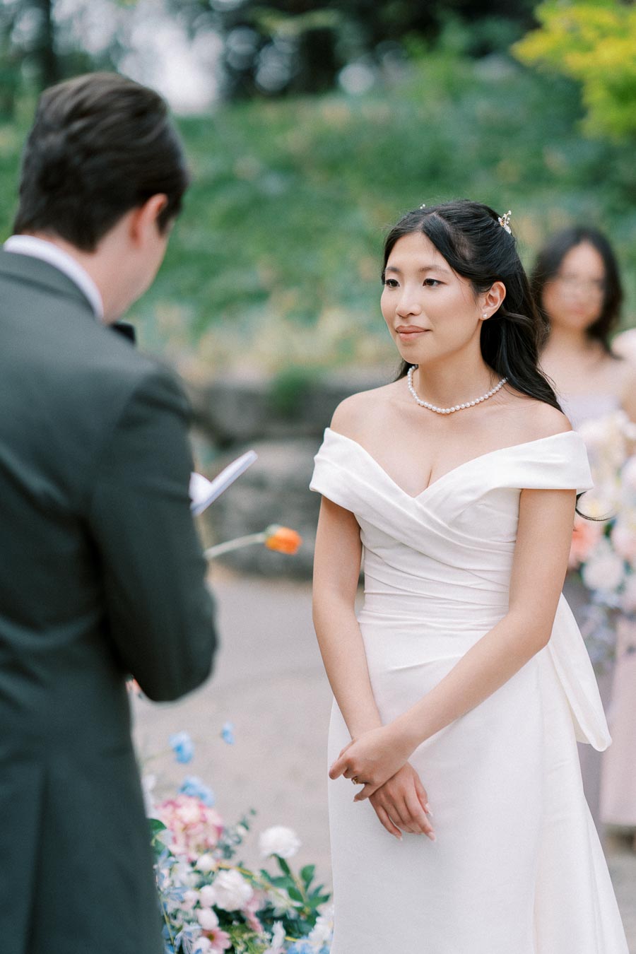 A bride in an elegant white wedding dress with off-the-shoulder design and a pearl necklace listens to vows during an outdoor ceremony, with a groom in a suit visible in the foreground and colorful flowers in the background.