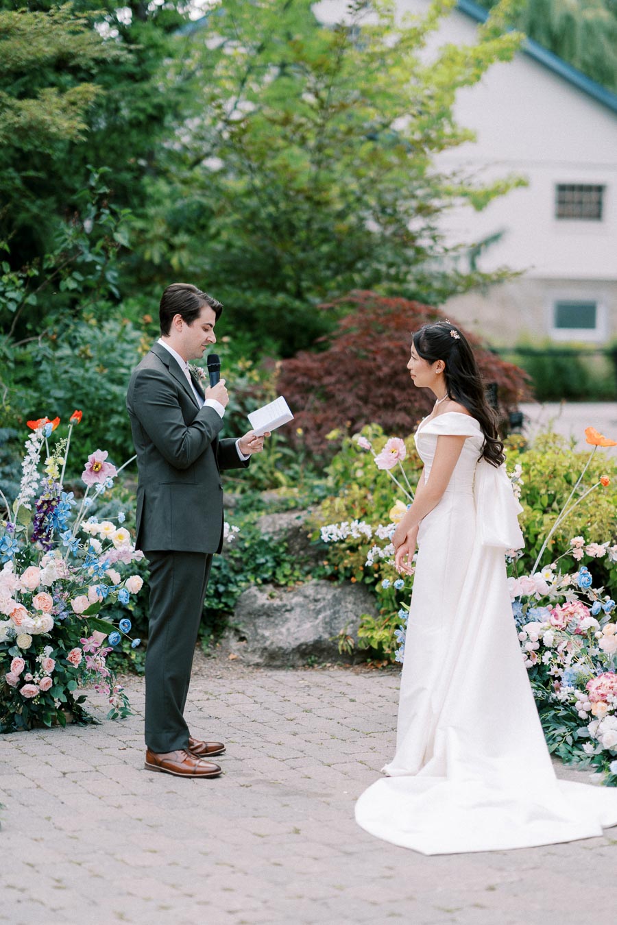 Wedding ceremony in a garden setting, with a groom in a suit reading vows to a bride in a white dress, surrounded by colorful floral arrangements.