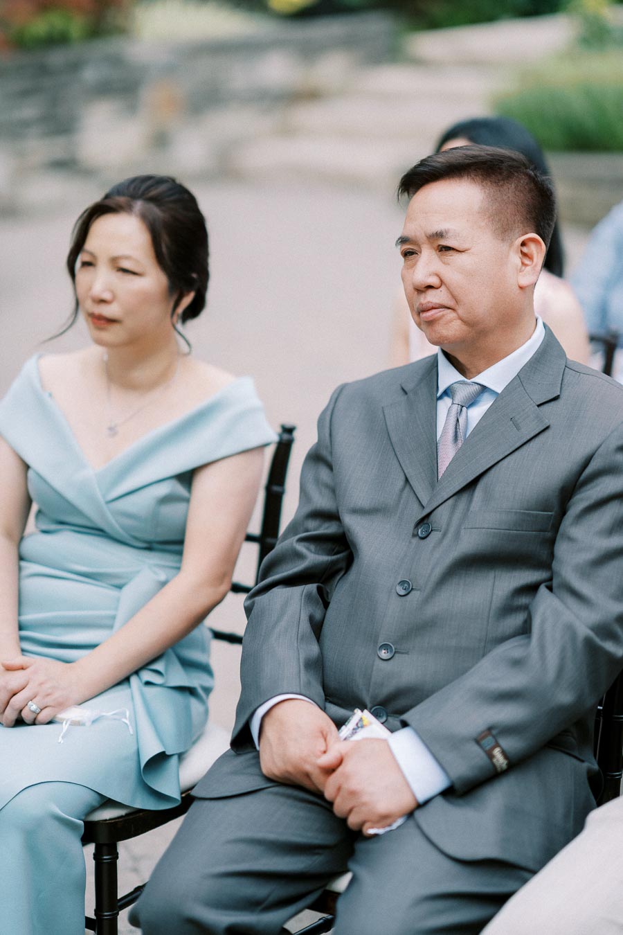 A couple sitting together wearing formal attire; a woman in a light blue dress and a man in a gray suit, attending an outdoor event with a blurred natural background.