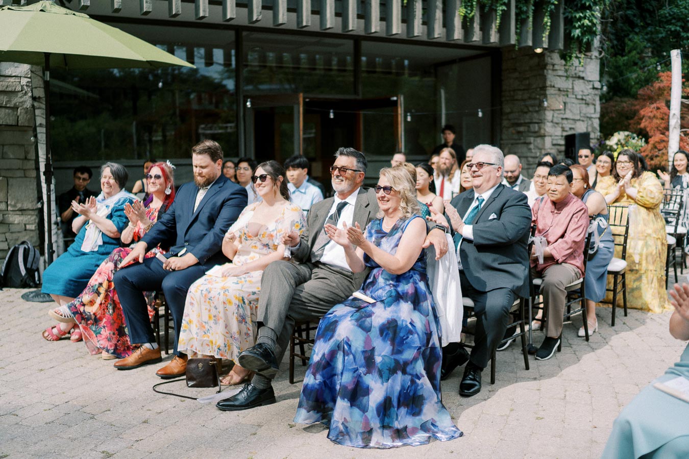 Audience at an outdoor event clapping, wearing colorful attire and sunglasses, seated in a sunny venue with stone and greenery decor.