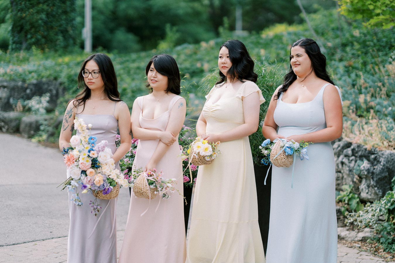 Four bridesmaids in pastel dresses holding floral arrangements at an outdoor wedding, surrounded by lush greenery.