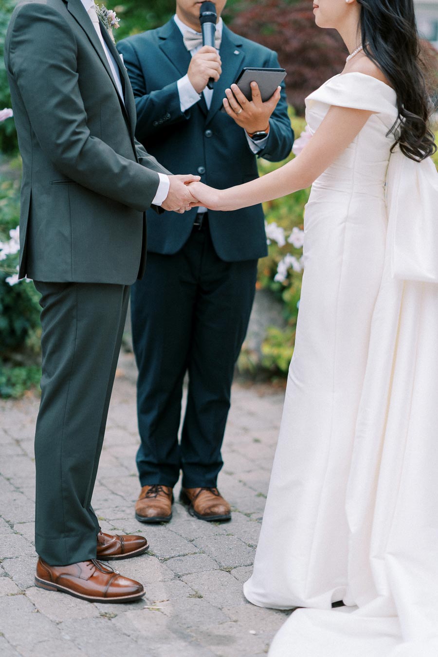 Wedding ceremony with a couple holding hands, an officiant standing nearby with a microphone, outdoors with greenery in the background.