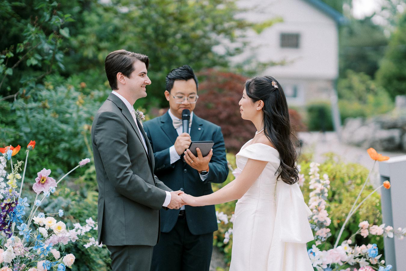 Outdoor wedding ceremony with couple holding hands and officiant reading vows, surrounded by colorful flowers.