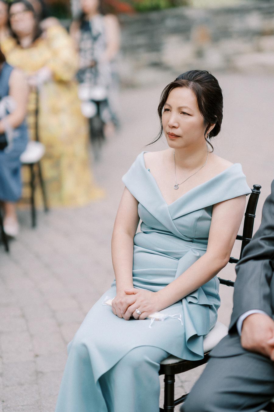 A woman in a light blue dress sits attentively on a chair at an outdoor event, surrounded by other attendees in a soft-focus background.