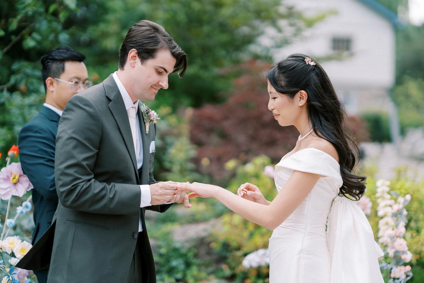 Bride and groom exchanging rings during an outdoor wedding ceremony, surrounded by lush greenery and colorful flowers.
