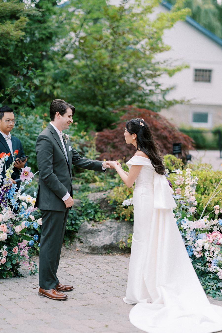 Outdoor wedding ceremony with bride in white dress and groom in gray suit exchanging vows amidst colorful floral arrangements.