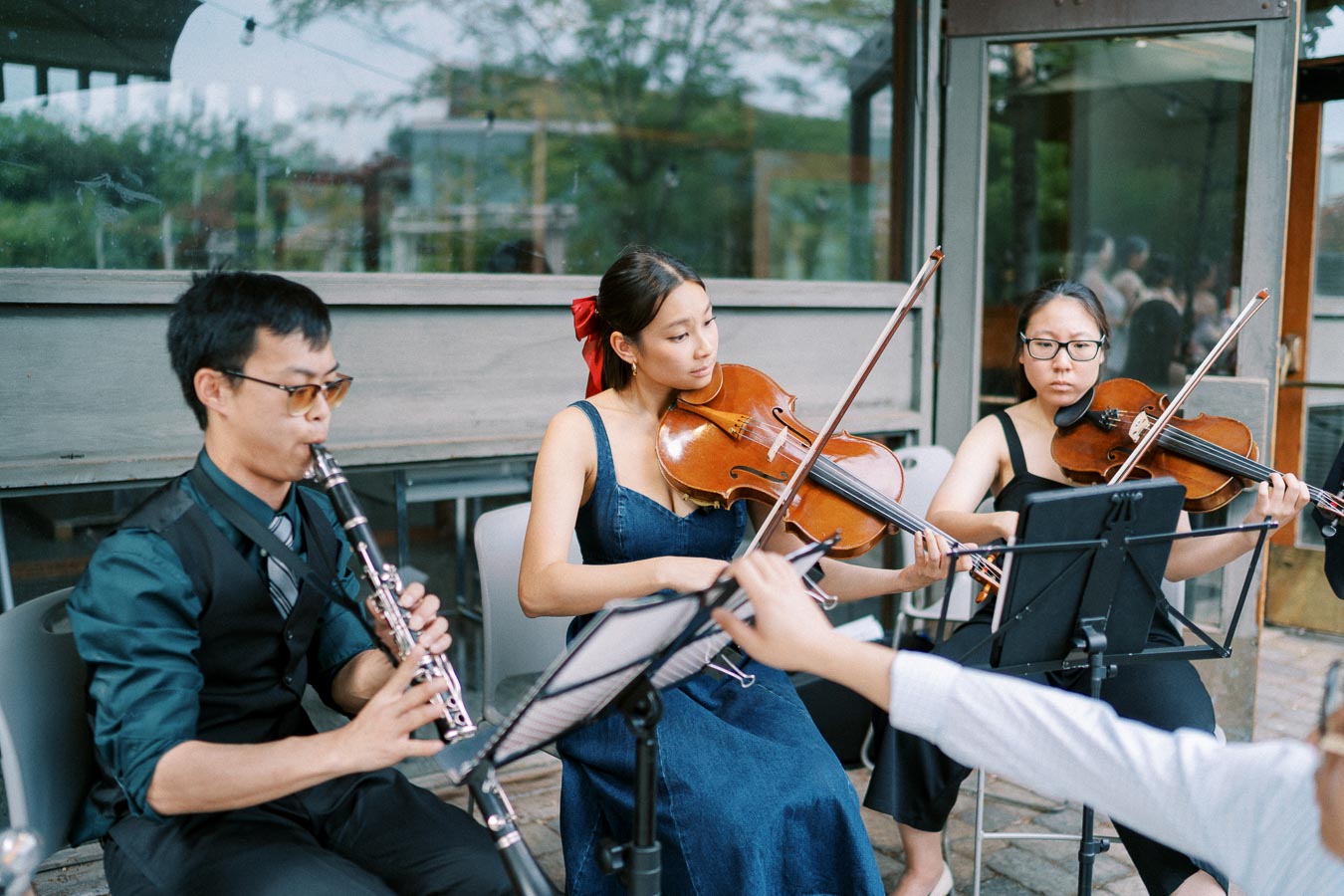 Outdoor musical ensemble with clarinet and violin players performing, featuring a woman in a blue dress with a red bow, creating a serene atmosphere.