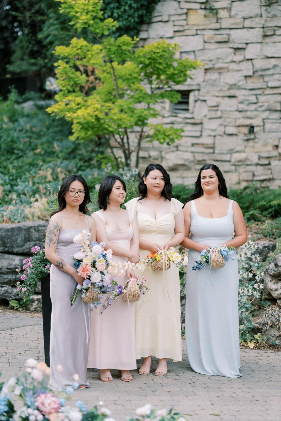 Bridesmaids in pastel dresses holding colorful bouquets, posing in a scenic garden setting with stone walls and lush greenery.