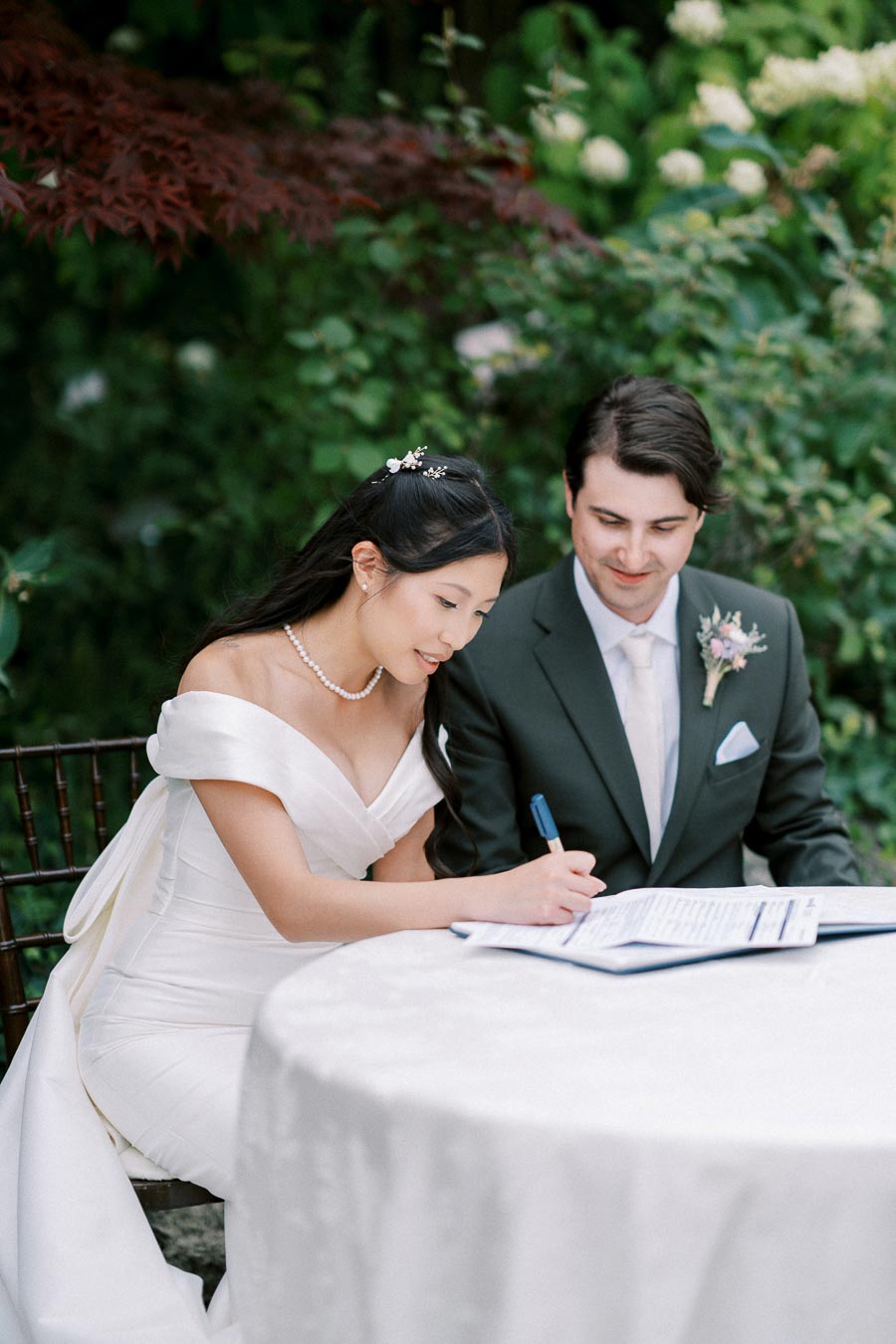 A bride and groom signing their marriage license at an outdoor wedding ceremony, surrounded by lush greenery and wearing elegant attire.