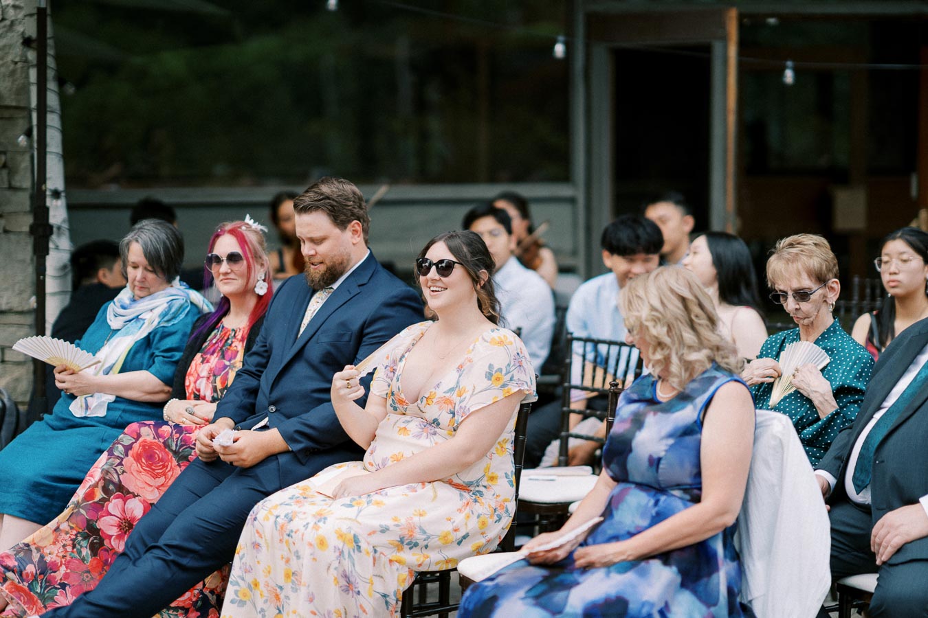 Group of people sitting outdoors at a wedding ceremony, dressed in colorful formal attire, with some using handheld fans to stay cool.
