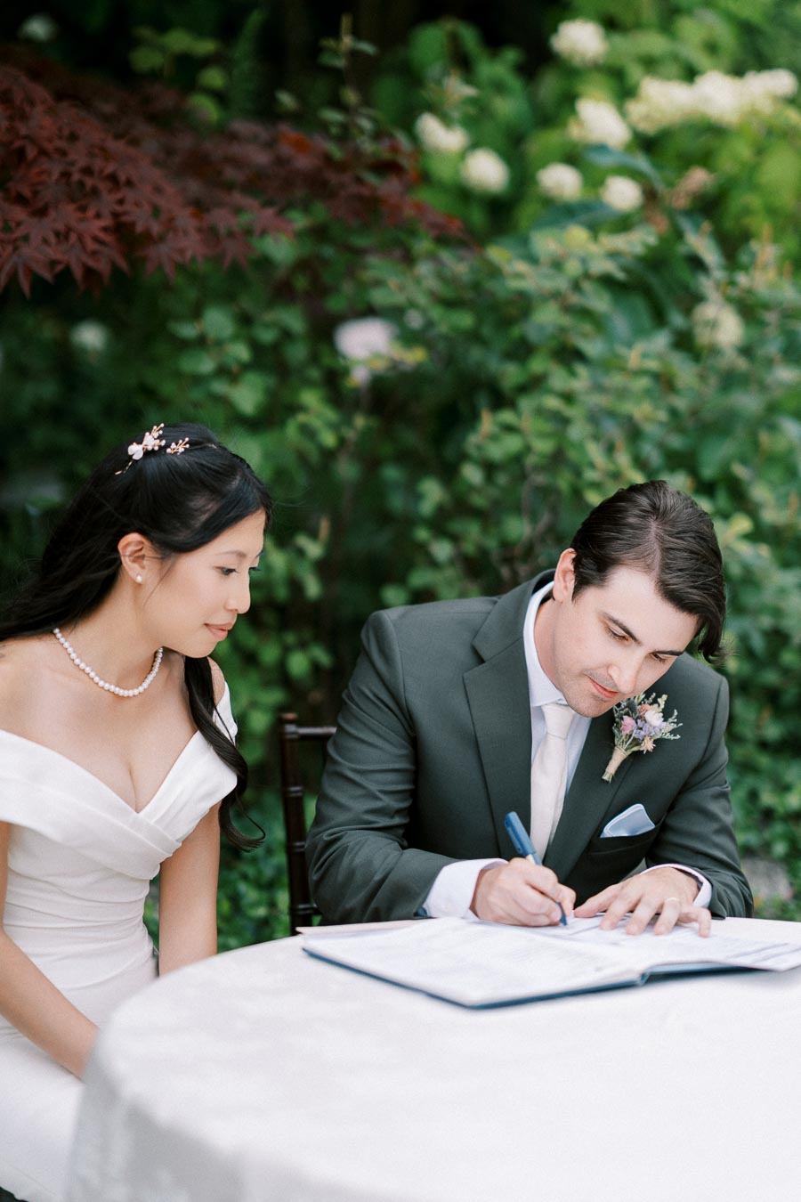 A bride and groom sign a marriage certificate at an outdoor wedding ceremony, surrounded by lush greenery. The bride is in an elegant off-shoulder white dress, while the groom wears a stylish dark suit with a floral boutonniere.