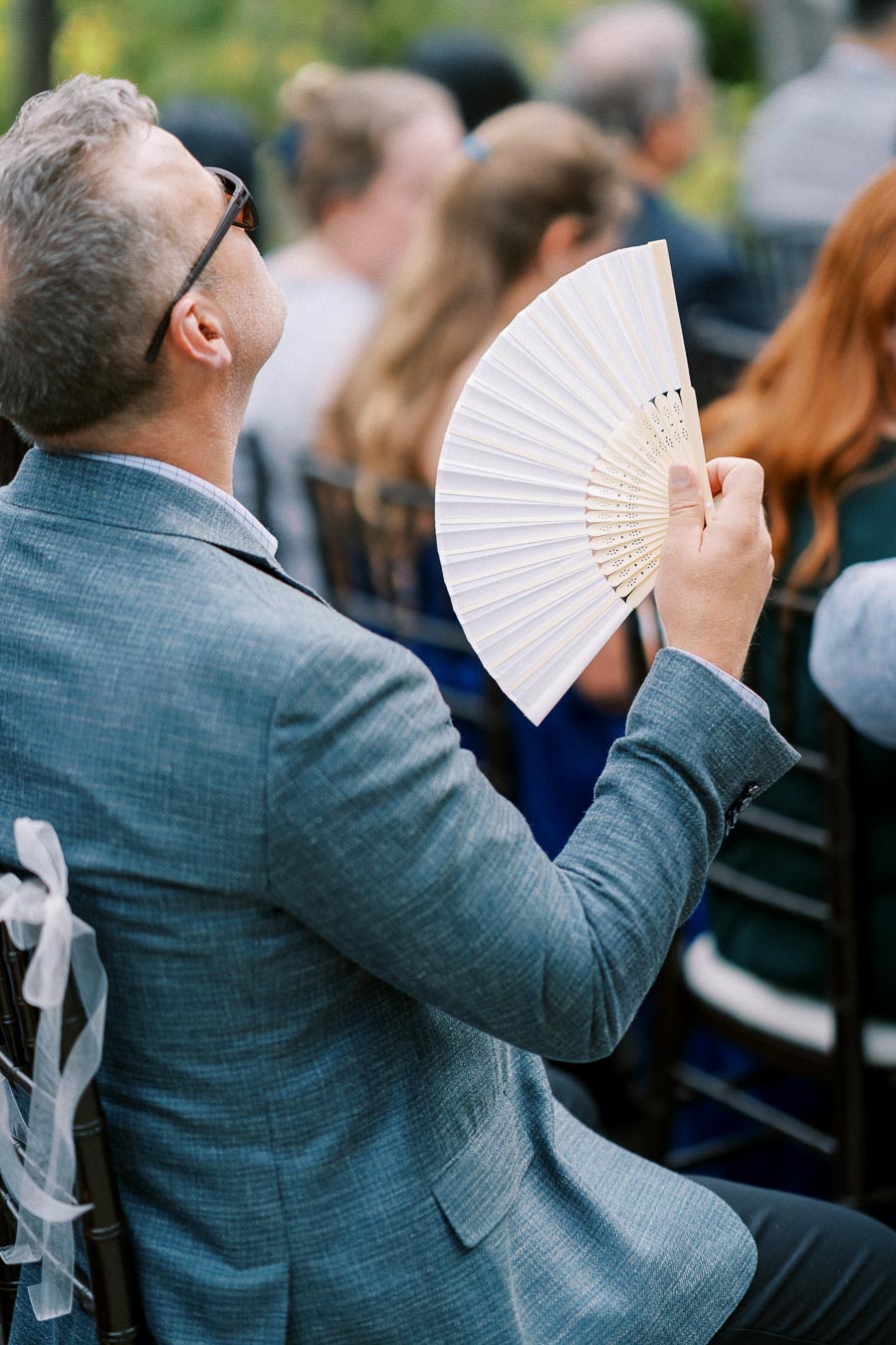 Man in gray suit holding a white hand fan at an outdoor event.
