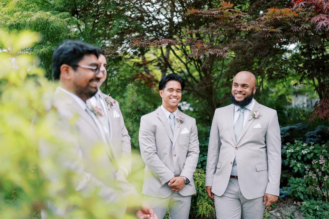 A group of groomsmen in light gray suits with botanical boutonnieres, standing outdoors surrounded by lush greenery and trees, smiling and engaging with each other.