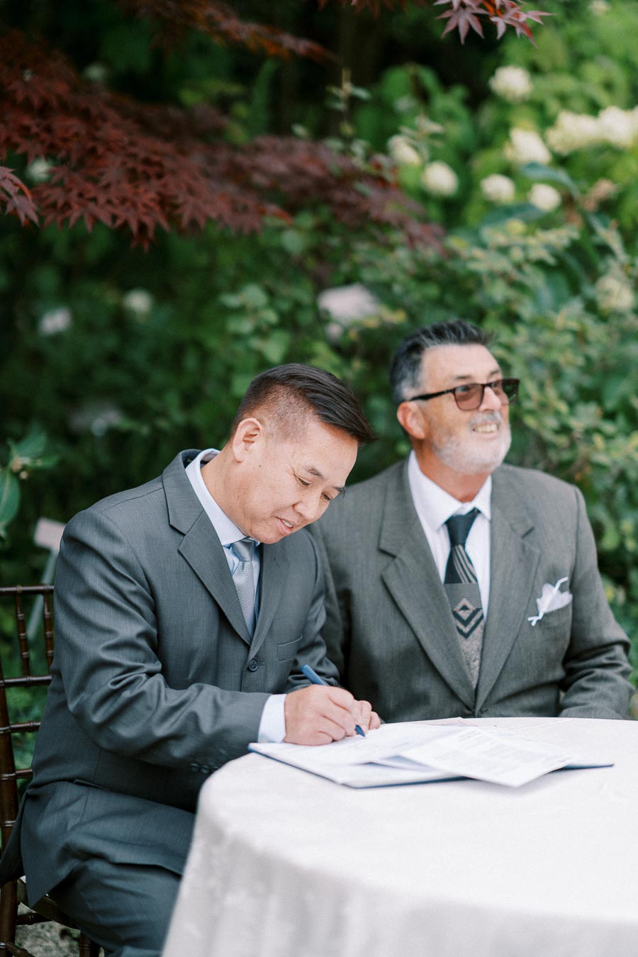 Two men in suits sitting outdoors at a table, one signing a document amidst a lush green garden backdrop.