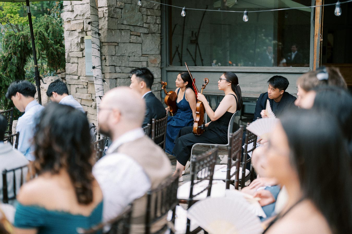 A small string ensemble sitting among guests at an outdoor event, some holding their violins and violas, with stone walls and greenery in the background.