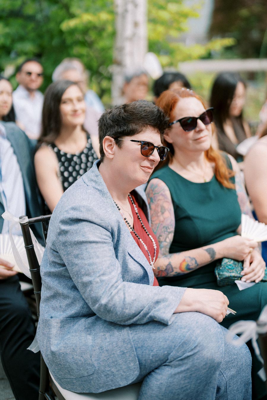 Two individuals sitting outdoors at a daytime event. One wears a light blue suit and sunglasses, the other a dark dress and sunglasses, displaying colorful tattoos. They appear relaxed and attentive, surrounded by other attendees.
