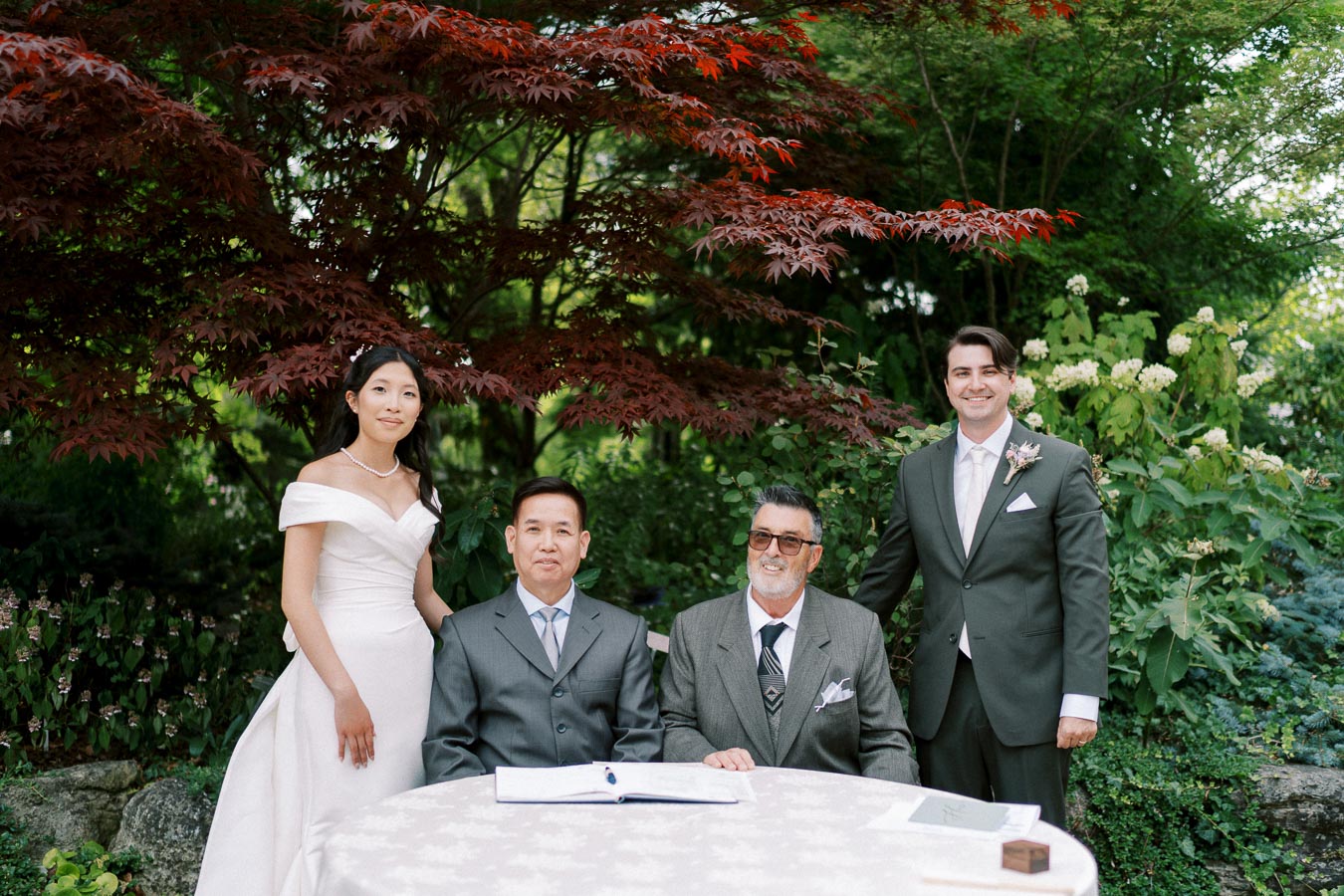 A bride and groom in formal attire pose with two older men at an outdoor wedding ceremony surrounded by lush greenery and a vibrant red tree.