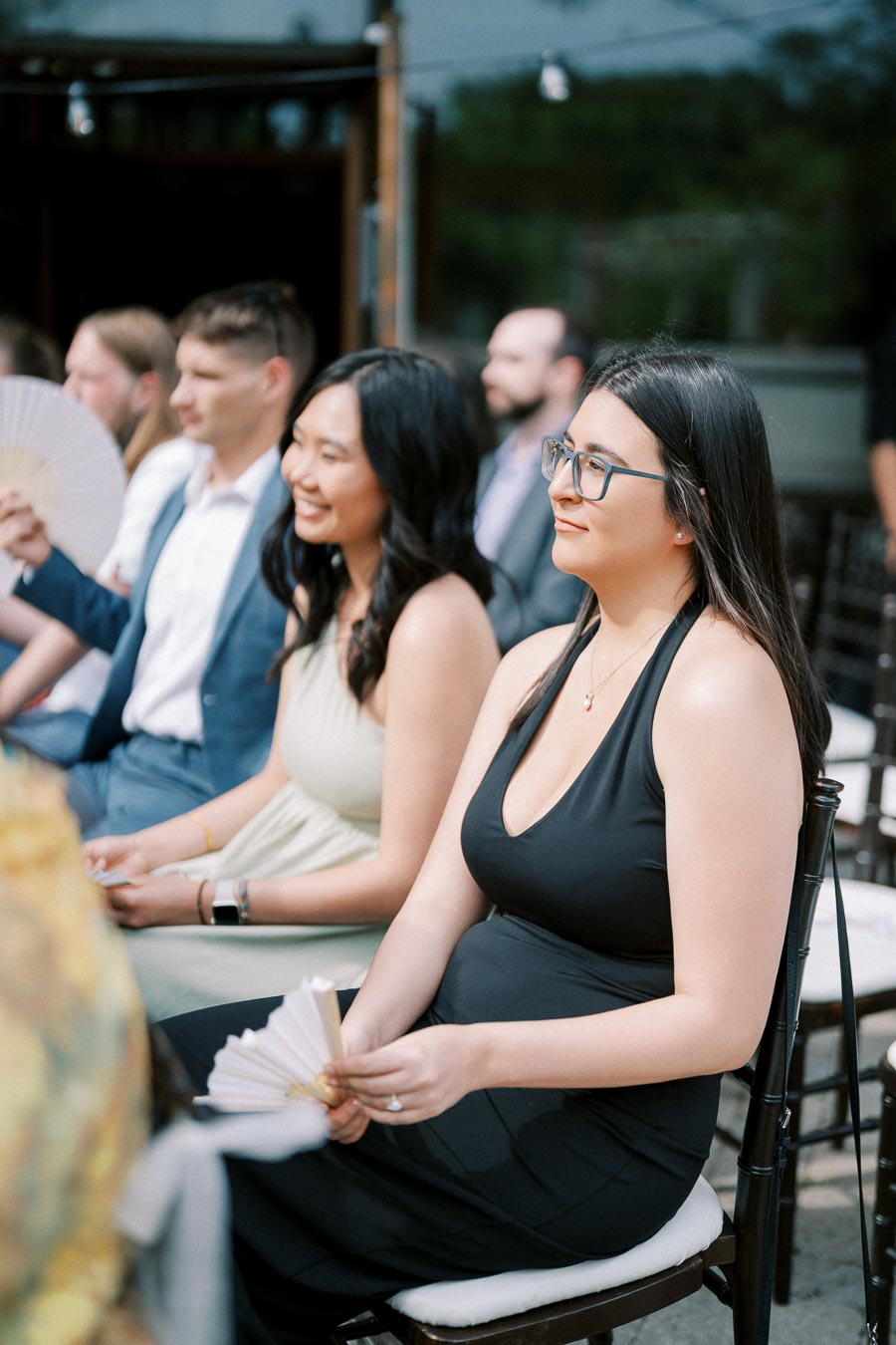 Guests seated at an outdoor event, enjoying the ceremony while using handheld fans, dressed in formal attire.
