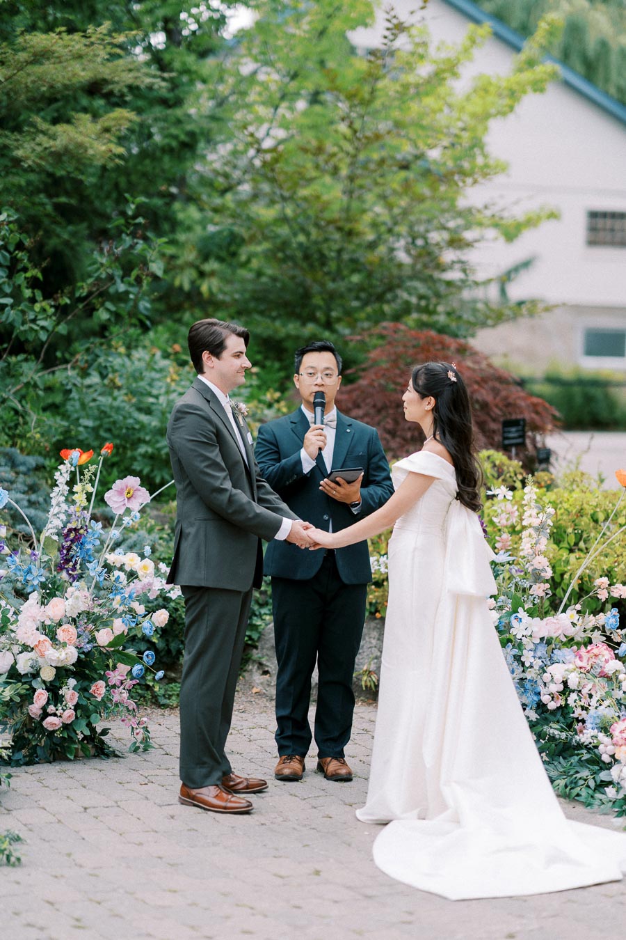 Outdoor wedding ceremony with a bride in a white gown and a groom in a suit exchanging vows, an officiant holding a microphone, surrounded by lush greenery and colorful floral arrangements.