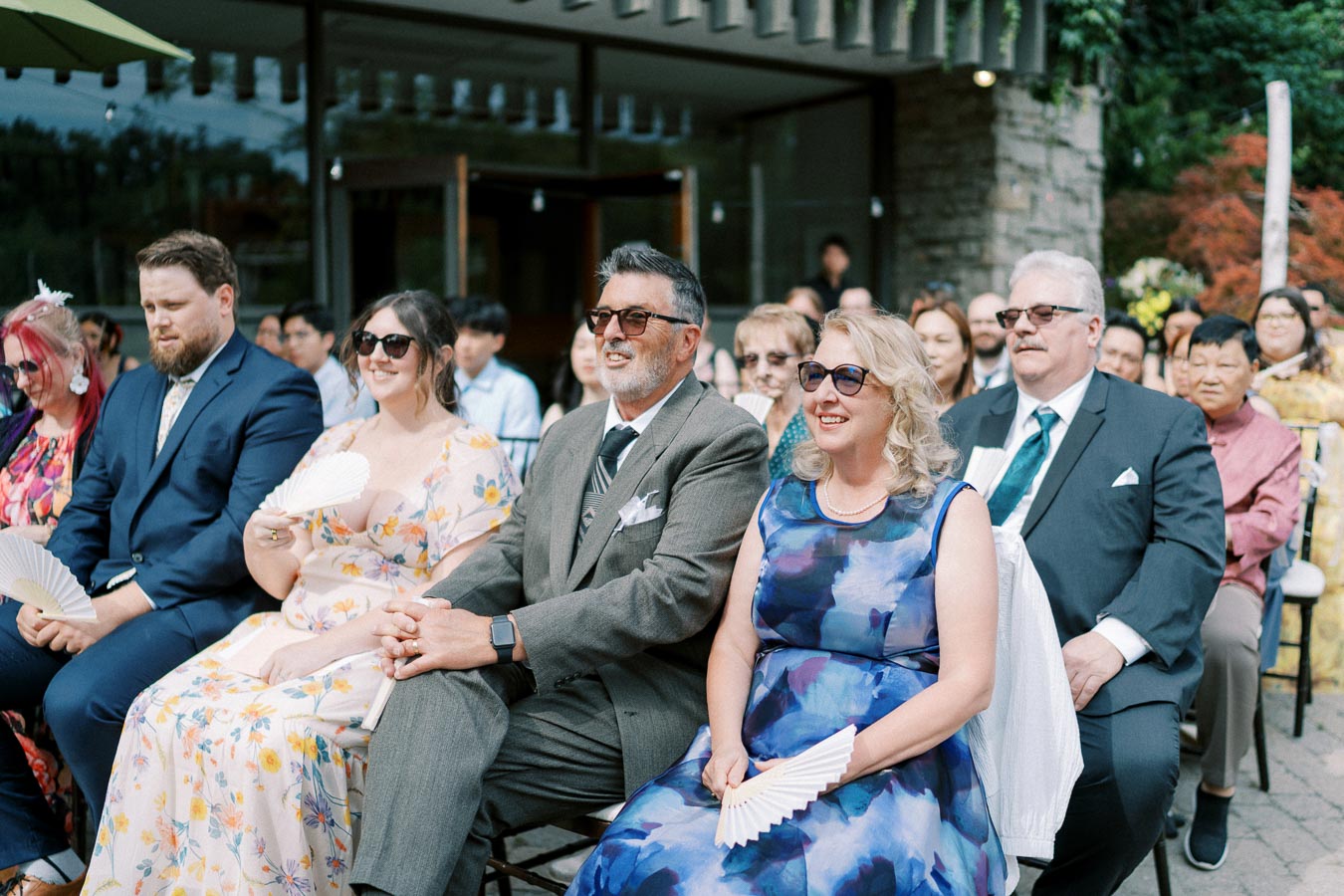 A group of well-dressed wedding guests seated outdoors, wearing sunglasses and holding hand fans, enjoying a sunny day at a wedding ceremony.