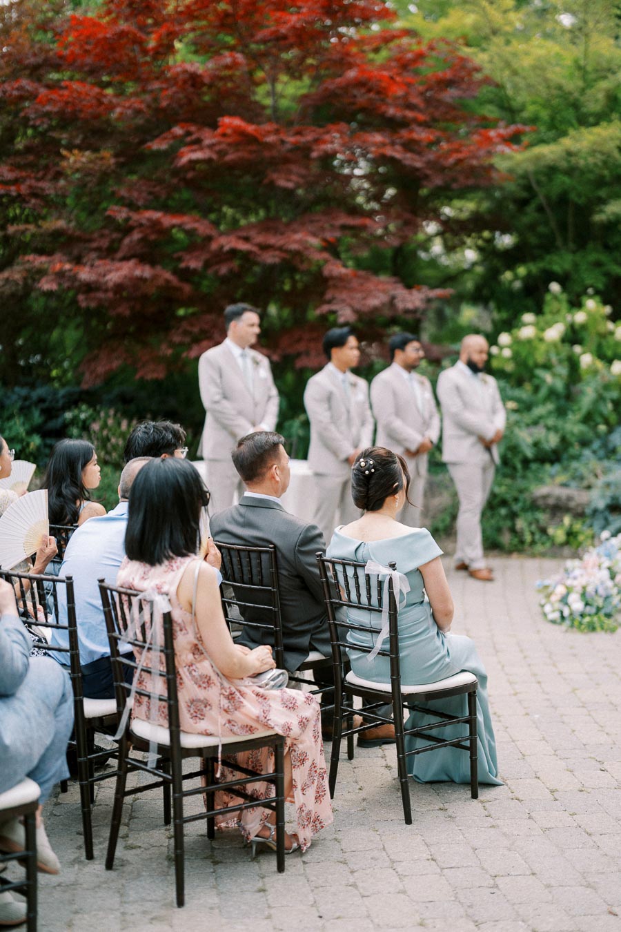 A wedding ceremony set outdoors with guests seated on wooden chairs, observing men in suits standing near lush greenery and vibrant red maple trees in the background.