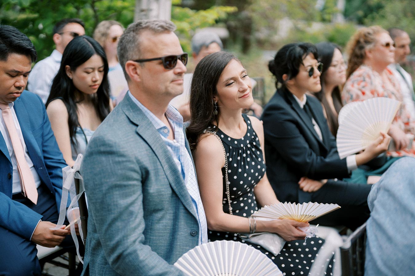 Audience at an outdoor wedding ceremony, dressed in formal attire and using fans to stay cool during a sunny day.