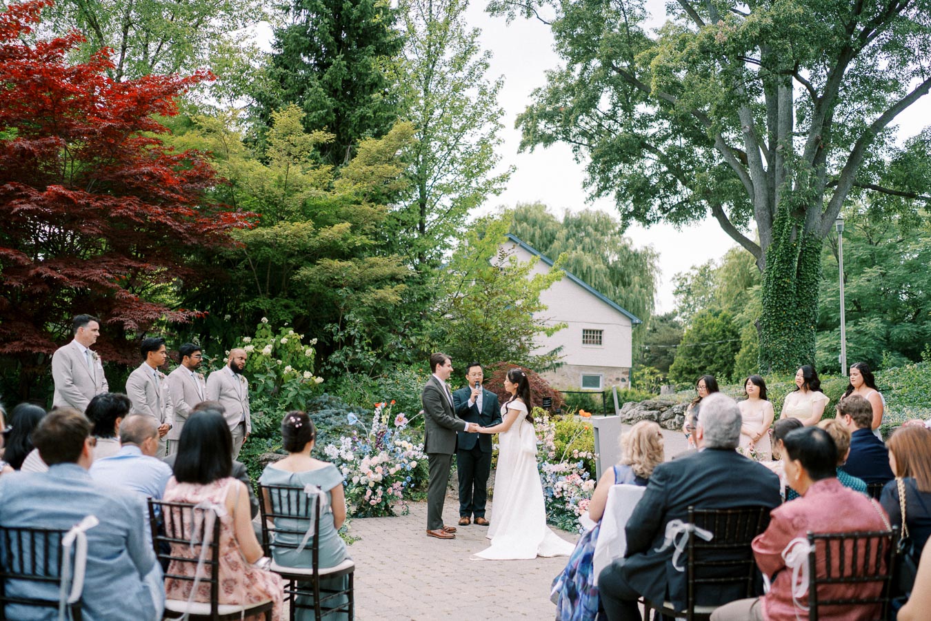 Outdoor garden wedding ceremony with a couple exchanging vows in front of an officiant, surrounded by colorful flowers and lush greenery, with guests seated on chairs in the foreground.