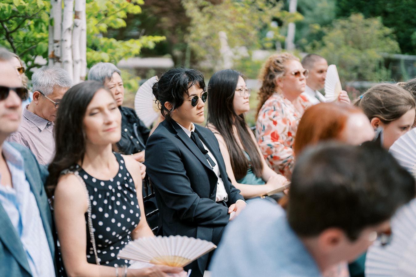 Group of people sitting outdoors at an event, holding fans and listening attentively, with greenery in the background.