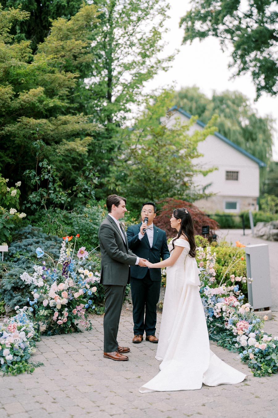 Outdoor wedding ceremony with a couple exchanging vows, surrounded by lush greenery and colorful floral arrangements. The bride wears a white gown, and the groom is dressed in a dark suit. A third person officiates the ceremony under a clear sky.