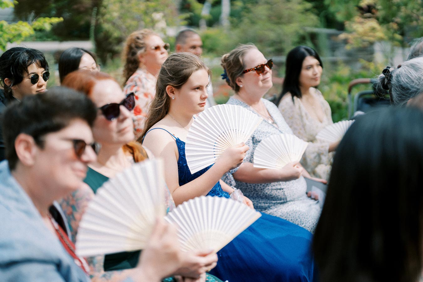 Group of people seated outdoors using hand fans, wearing summer attire and sunglasses, enjoying a sunny day.