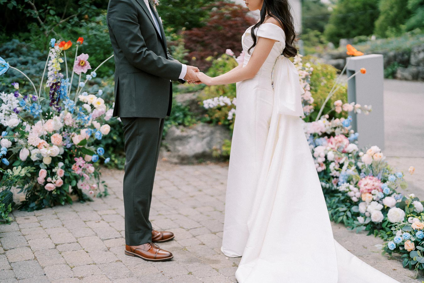 A bride and groom holding hands during an outdoor wedding ceremony, surrounded by colorful floral arrangements on a stone pathway.