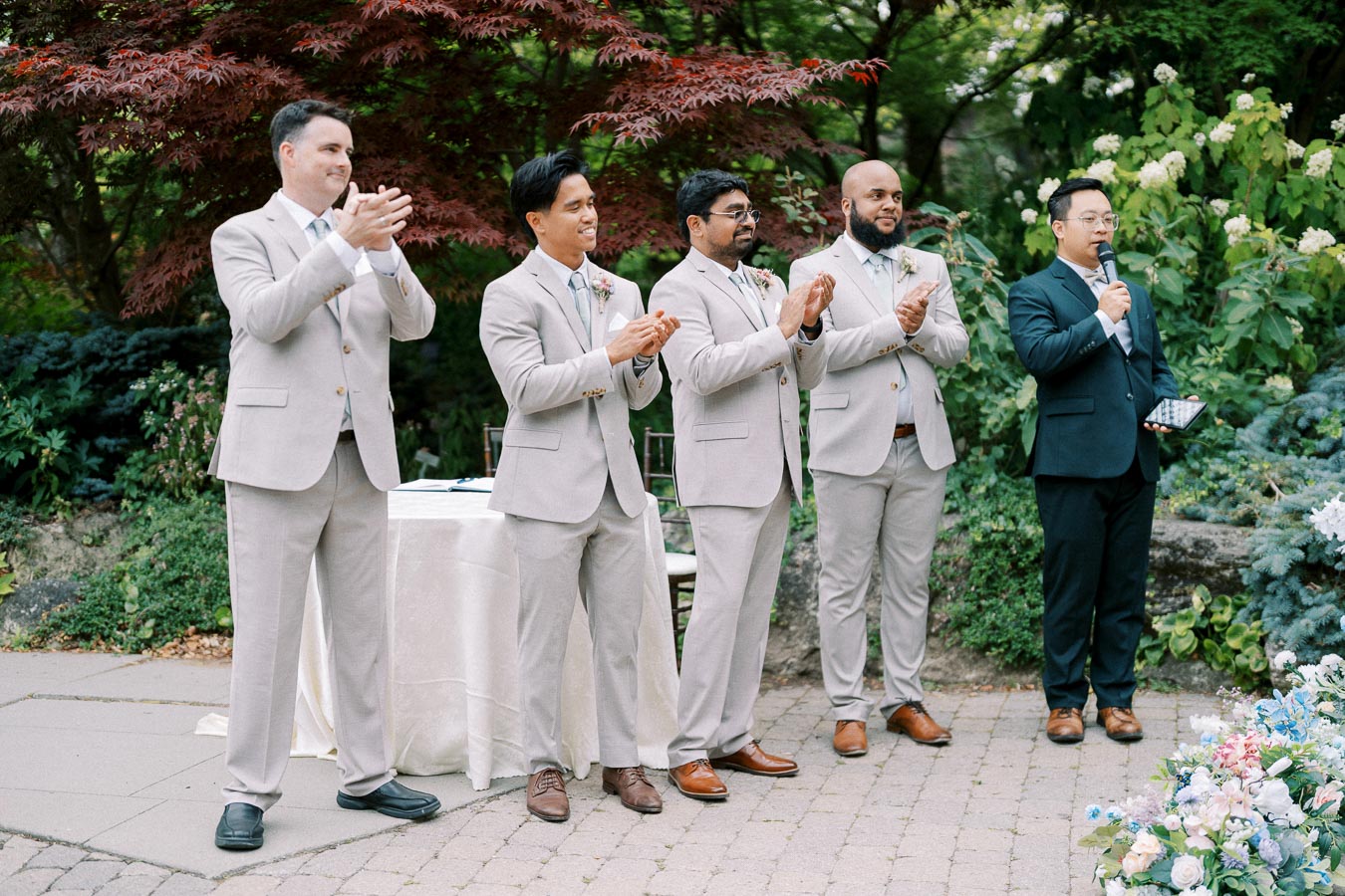 Groomsmen in beige suits clapping at an outdoor wedding ceremony with lush greenery and flowers.