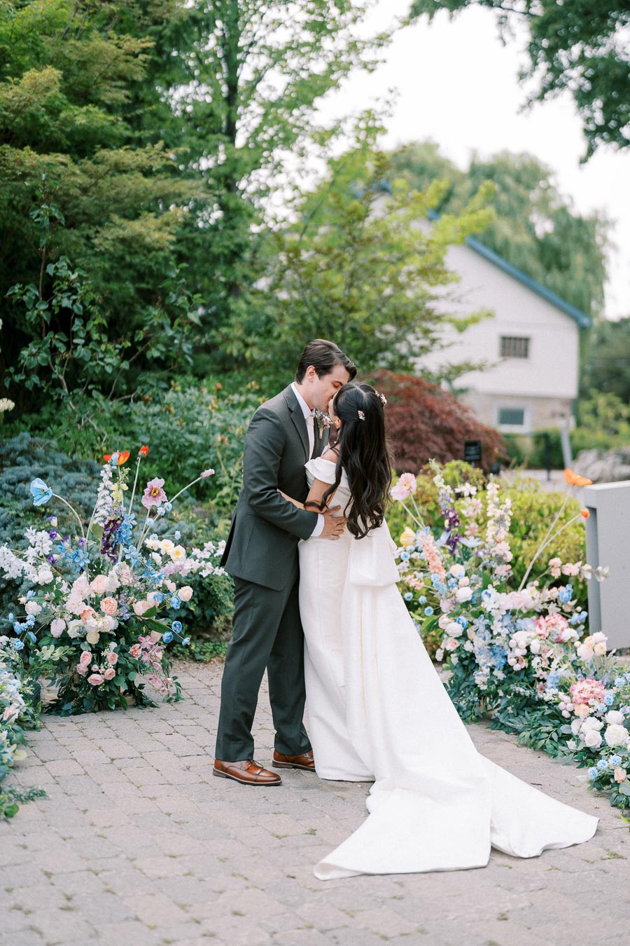 A couple shares a romantic kiss during their outdoor wedding ceremony, surrounded by vibrant floral arrangements and lush greenery.