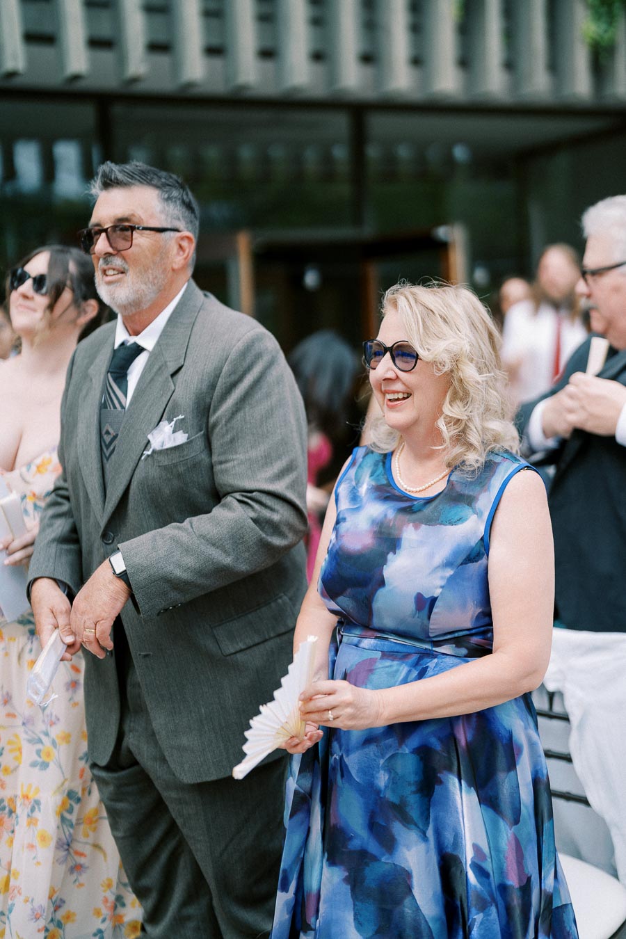 Elderly couple smiling at an outdoor event, with the man in a suit and the woman in a blue floral dress holding a paper fan, surrounded by casually dressed attendees.