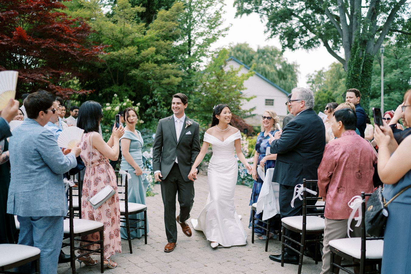 Wedding ceremony with a bride in a white dress and a groom in a suit walking down an outdoor aisle, surrounded by smiling guests capturing the moment on their phones amidst lush greenery and decorative elements.