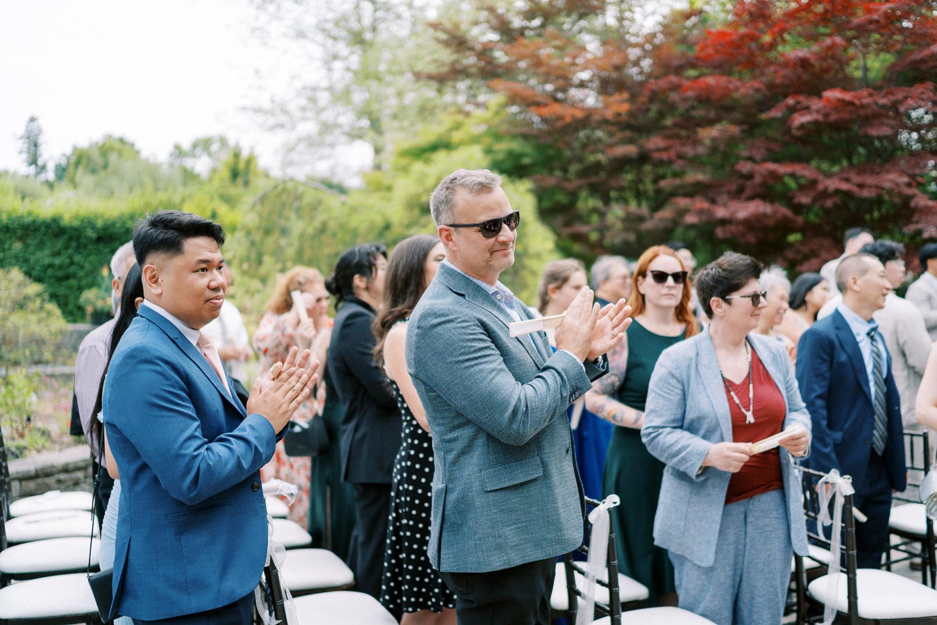 People standing and clapping at an outdoor event, surrounded by lush greenery and vibrant red trees.