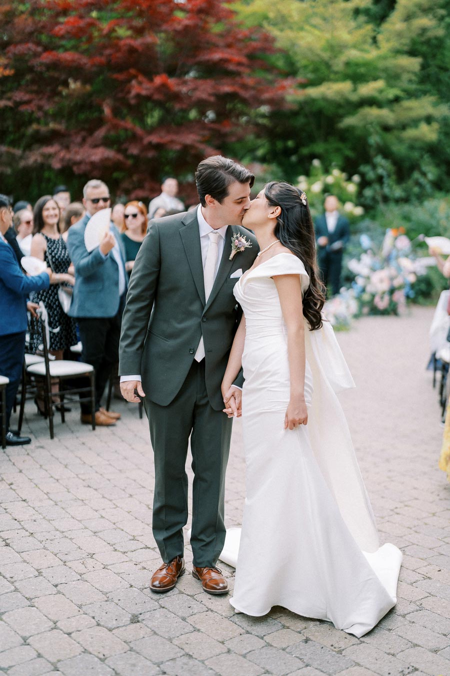 Newlywed couple shares a romantic kiss outdoors, surrounded by wedding guests, vibrant greenery, and colorful flowers, capturing a beautiful and joyous moment.